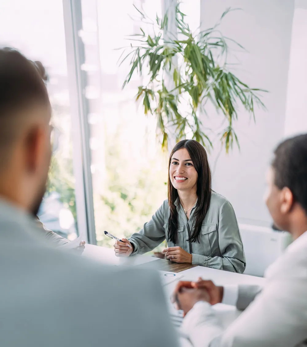 Junge Geschäftsfrau sitzt lächelnd in einem modernen Besprechungsraum und spricht mit Kolleginnen und Kollegen bei einem Meeting. Helle Fensterfronten und Pflanzen schaffen eine freundliche Arbeitsatmosphäre.
